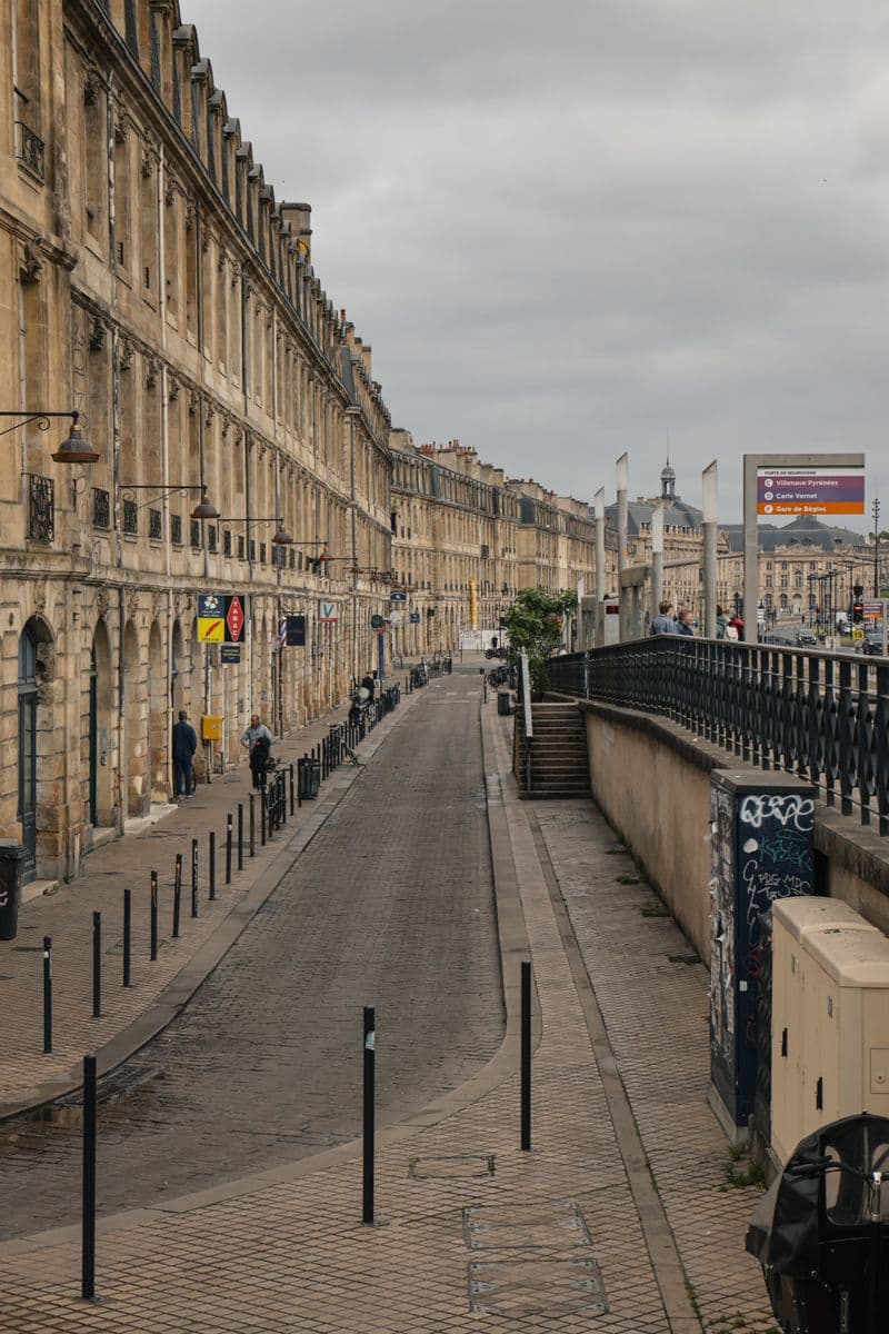 Rue pavée de Bordeaux bordée de façades en pierre et de potelets, sous un ciel couvert